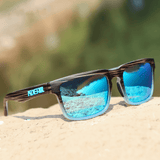 Óculos de sol Tiger modelo dia a dia em ângulo diagonal sob a areia com paisagem ao fundo na cor marrom com azul, disponível em: ethosloja.com.br